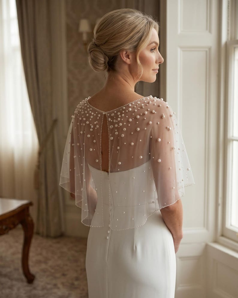 K&ouml;nnte beinhalten: Ein wei&szlig;es &auml;rmelloses Kleid mit einem transparenten T&uuml;ll-Capelet, das mit verstreuten wei&szlig;en Perlen unterschiedlicher Gr&ouml;&szlig;e verziert ist. Das Capelet hat einen runden Ausschnitt, eine Schl&uuml;sselloch-&Ouml;ffnung am R&uuml;cken und einen perlenbesetzten Rand.