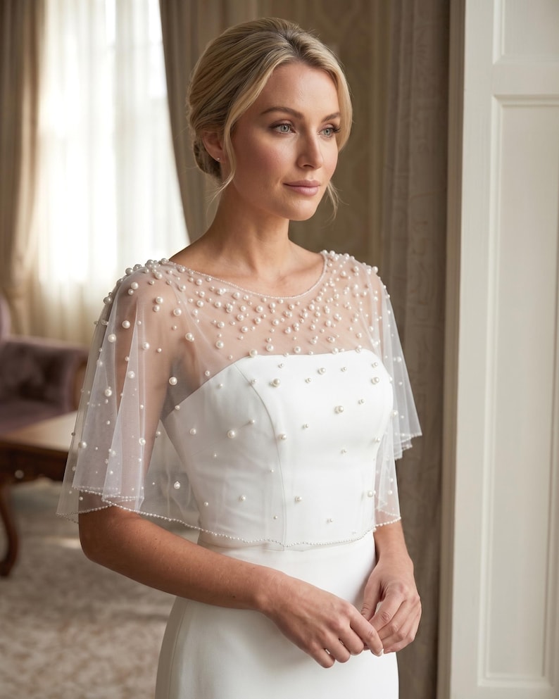 K&ouml;nnte beinhalten: Ein wei&szlig;es Braut-Capelet mit einem transparenten T&uuml;ll&uuml;berwurf, der mit verstreuten wei&szlig;en Perlen unterschiedlicher Gr&ouml;&szlig;e verziert ist. Das Capelet hat einen runden Ausschnitt und kurze, flie&szlig;ende &Auml;rmel, die &uuml;ber einem schlichten wei&szlig;en Kleid liegen.