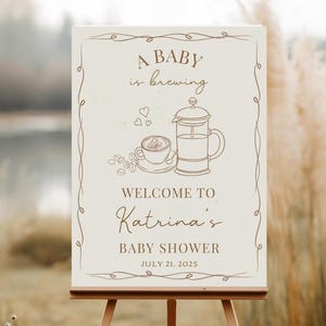 May include: A cream-colored sign on a wooden easel with the text "A BABY is brewing" and "WELCOME TO Katrina's BABY SHOWER JULY 21, 2025". The sign features a coffee-themed illustration with a French press and a cup of coffee.