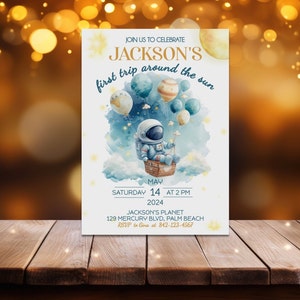 May include: A white invitation card with a blue watercolor background featuring an astronaut in a hot air balloon with planets and stars. The text reads "JOIN US TO CELEBRATE JACKSON'S first trip around the sun MAY 14 AT 2PM 2024 JACKSON'S PLANET 129 MERCURY BLVD, PALM BEACH RSVP to Give at 842-123-4567"