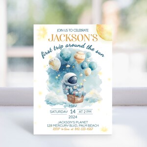 May include: A blue and white astronaut-themed first birthday party invitation with a watercolor background. The invitation features a cartoon astronaut holding a star in a basket attached to balloons. The text reads "Join us to celebrate Jackson's first trip around the sun" and includes the date, time, and location of the party.