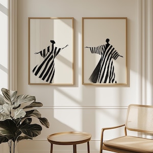 May include: Two framed art prints featuring black and white striped figures in flowing robes. The figures are set against a cream-colored background, with a simple wooden frame. The artwork is displayed on a white wall.
