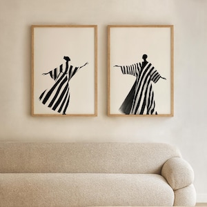 May include: Two framed art prints featuring black and white striped figures in flowing garments. The figures are set against a cream background, with light brown wooden frames. The artwork is displayed above a beige sofa and a wooden coffee table.