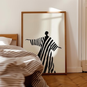 May include: A framed art print featuring a black and white striped figure with outstretched arms. The artwork is set against a cream background and framed in a natural wood. The print is displayed in a bedroom setting.