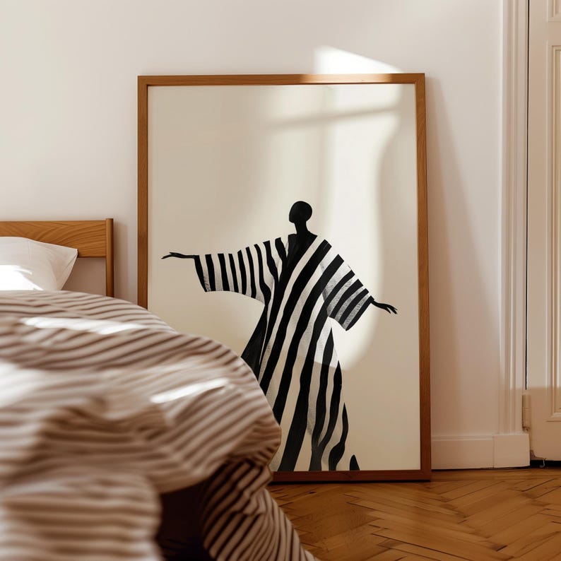 May include: A framed art print featuring a black and white striped figure with outstretched arms. The artwork is set against a cream background and framed in a natural wood. The print is displayed in a bedroom setting.