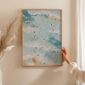 May include: An abstract painting of surfers in the ocean. The artwork features a light blue and white color palette, with small figures on surfboards scattered across the canvas. The painting is framed in a light brown wooden frame.