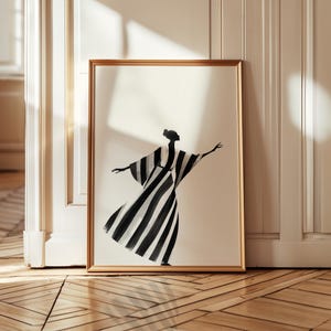 May include: A framed art print featuring a black and white striped figure in a flowing dress with arms outstretched. The artwork is set against a cream background within a gold-colored frame. The print is displayed against a white wall and a wooden floor.