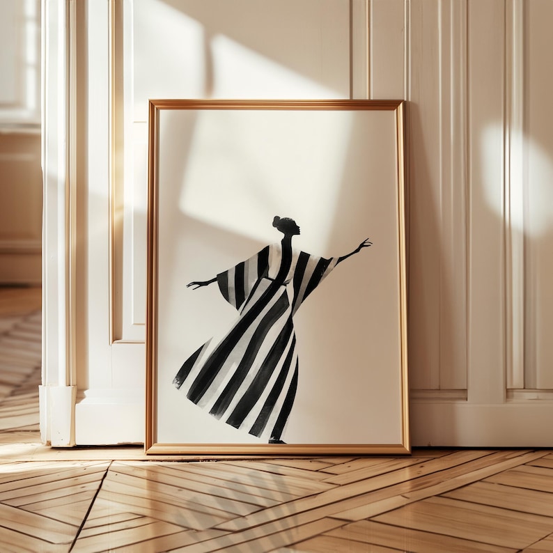 May include: A framed art print featuring a black and white striped figure in a flowing dress with arms outstretched. The artwork is set against a cream background within a gold-colored frame. The print is displayed against a white wall and a wooden floor.