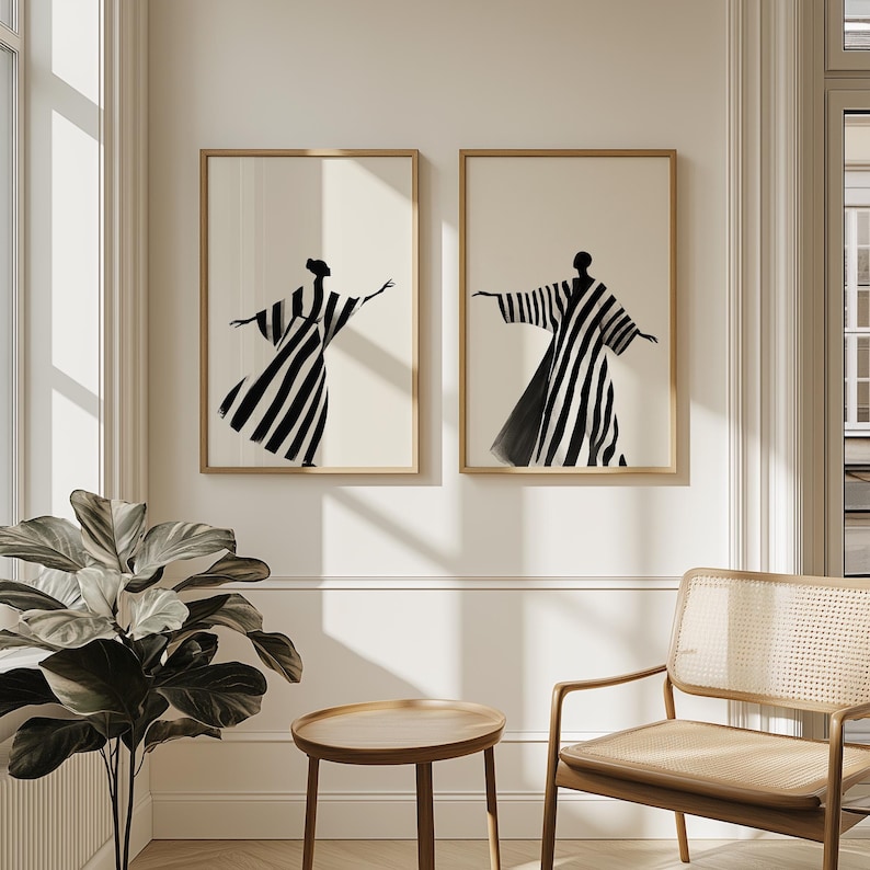 May include: Two framed art prints featuring black and white striped figures in flowing robes. The figures are set against a cream-colored background, with a simple wooden frame. The artwork is displayed on a white wall.