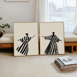 May include: Two framed art prints featuring abstract figures in black and white striped robes. The figures are set against a cream-colored background, with gold-colored frames. The artwork is displayed on a light-colored carpet.