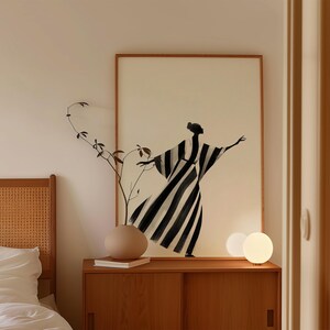 May include: A framed art print featuring a black and white striped figure with outstretched arms. The artwork hangs above a wooden cabinet with a vase and a sprig of greenery. Two spherical lamps sit on the cabinet.