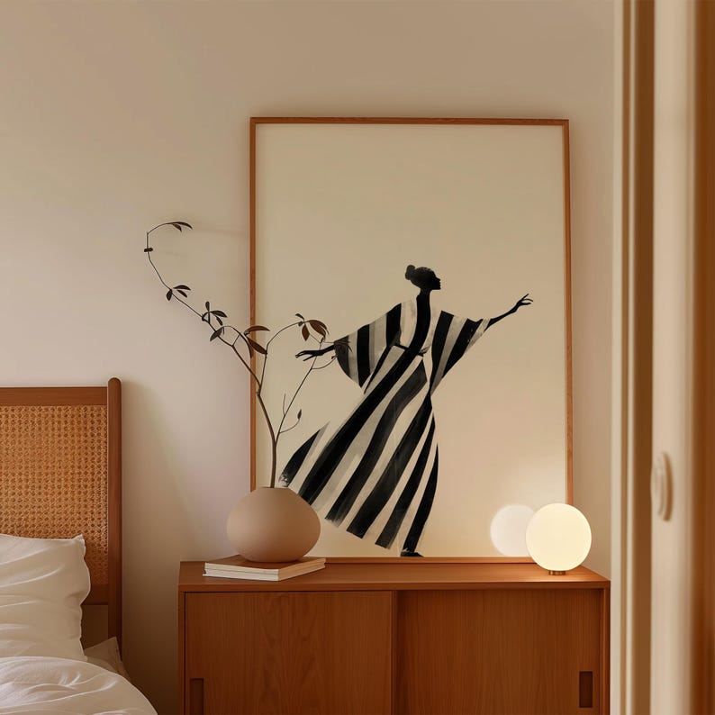 May include: A framed art print featuring a black and white striped figure with outstretched arms. The artwork hangs above a wooden cabinet with a vase and a sprig of greenery. Two spherical lamps sit on the cabinet.