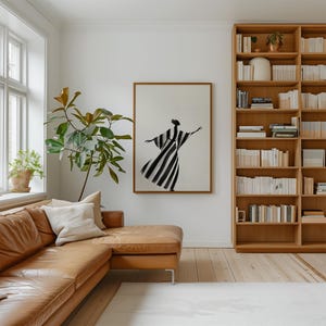 May include: A living room scene featuring a brown leather sectional sofa, a large wooden bookshelf filled with books, and a framed artwork depicting a figure in a black and white striped dress. A large plant and a window are also visible.