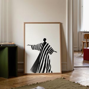 May include: A framed art print featuring a black and white illustration of a figure in a striped robe with outstretched arms. The artwork is set against a white background, framed in a light brown wooden frame. The print is displayed on a wooden floor.