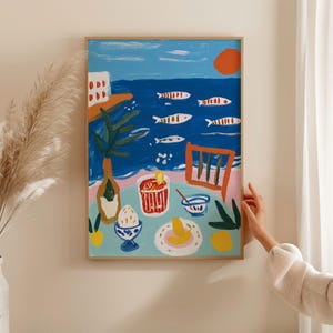 May include: A framed art print featuring a vibrant coastal scene with a blue sea, orange sun, and white fish. The foreground shows a table with a red drink, a bowl, a yellow fruit, and an egg in a blue cup, all rendered in a bold, painterly style.