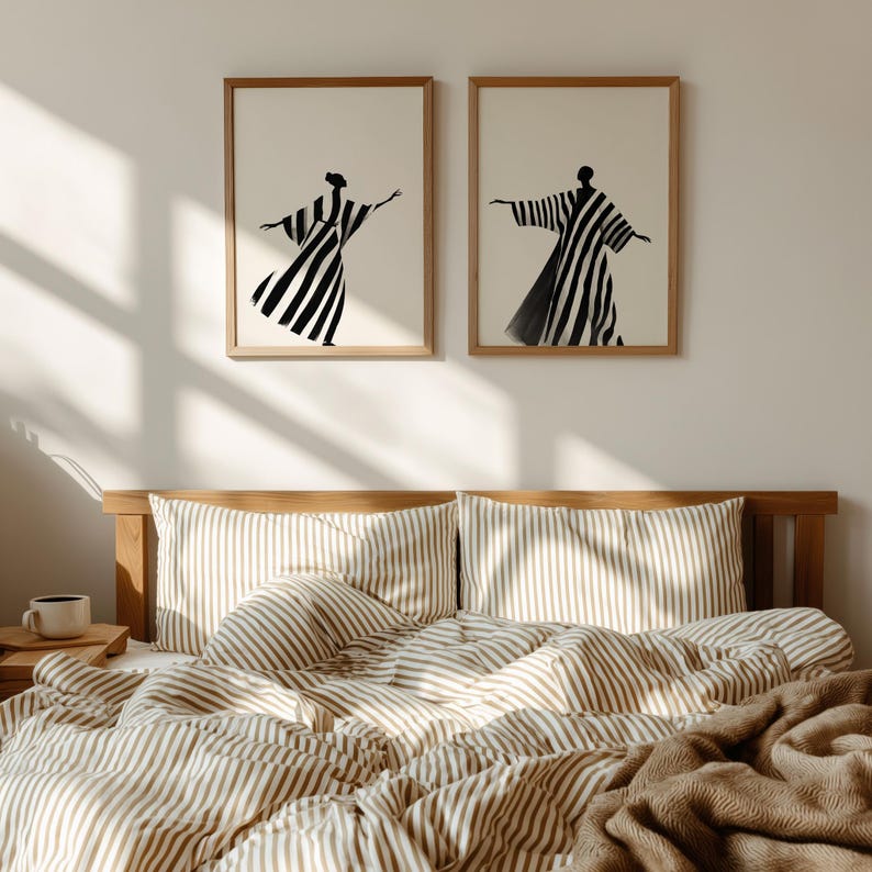 May include: Two framed art prints featuring abstract figures in black and white striped robes. The artwork hangs above a bed with striped bedding in shades of beige and white. The room has a minimalist, modern aesthetic.