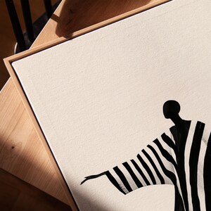 May include: Abstract art print featuring a figure in a black and white striped robe. The artwork is set against a cream-colored canvas, framed in natural wood. The figure's arm is outstretched, creating a sense of movement.