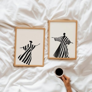 May include: Two framed art prints featuring abstract figures in black and white striped robes with outstretched arms. The prints are in light wood frames, set against a white, textured background. A hand holds a cup of coffee.