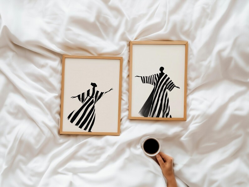 May include: Two framed art prints featuring abstract figures in black and white striped robes with outstretched arms. The prints are in light wood frames, set against a white, textured background. A hand holds a cup of coffee.