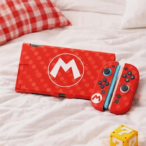 May include: Red Nintendo Switch case and controllers featuring a white "M" logo and star pattern. The controllers have black buttons and blue accents. A yellow question mark block is in the foreground.