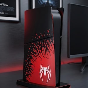 Op de afbeelding: Zwarte en rode gameconsole met een wit spinnenlogo. De console heeft een rode accentkleur en een zwarte basis. Het ontwerp heeft een rode achtergrond met een zwart, webachtig patroon. De console staat op een zwarte standaard.