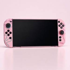 Może przedstawiać: Różowa konsola do gier przenośna z czarnym ekranem. Konsola ma kolorowe abstrakcyjne wzory i napis "UMMP" wydrukowany z boku. Przyciski i joysticki są czarne i różowe.