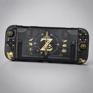 Puede incluir: Una consola de juegos portátil negra con estampados gráficos de color dorado. El diseño incluye un emblema central de una espada y la letra Z, rodeado de varios iconos pequeños, líneas intrincadas y siluetas de personajes en los mandos.