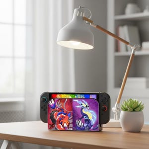 Könnte beinhalten: Eine schwarze Nintendo Switch-Konsole ist in einem Ständer mit zwei Zeichentrickfiguren angedockt. Eine weiße Schreibtischlampe mit Holzarm befindet sich über der Konsole. Rechts befindet sich eine kleine Topf-Sukkulente.