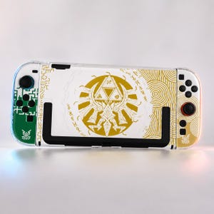May include: A white and gold Nintendo Switch case with a Triforce design. The case has a clear protective cover and green and black accents on the left side. The right side has black buttons and a red joystick.