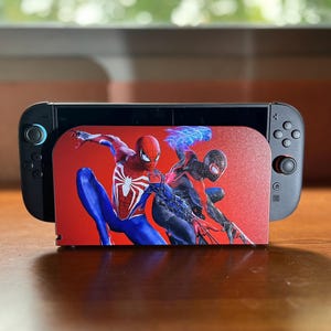 Può includere: Console Nintendo Switch nera e grigia con grafica di Spider-Man rossa e bianca sul retro. La console è appoggiata su una superficie marrone, con uno sfondo verde sfocato.