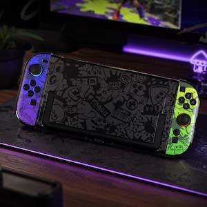 Peut inclure: Une console de jeu portable avec une unité centrale noire ornée d'un motif graffiti gris foncé. La manette gauche est violette avec des boutons noirs, et la manette droite est vert néon avec des boutons noirs et un anneau rouge autour du joystick.