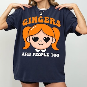 May include: Navy blue t-shirt with the text "GINGERS ARE PEOPLE TOO" in orange and white. The shirt features a cartoon illustration of a girl with orange pigtails and heart-shaped eyes.