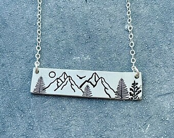Puede incluir: Un collar de barra plateada con un diseño de paisaje grabado en negro. El colgante muestra una cordillera, pinos, un pequeño sol y un pájaro en vuelo. La barra está suspendida de una delicada cadena de plata con eslabones circulares.