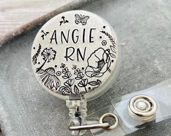 Puede incluir: Un portatarjetas circular con una cara de tono plateado cepillado que presenta diseños florales negros grabados, una mariposa y el texto ANGIE RN. El dispositivo incluye una correa de plástico transparente con un broche metálico a presión.