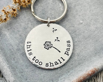 Puede incluir: Llavero de plata con un dije redondo que dice "this too shall pass" con un diseño de diente de león.