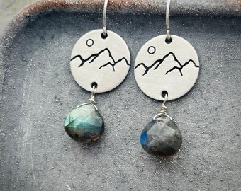 Puede incluir: Un par de pendientes colgantes circulares de tono plateado con un diseño estampado de montañas y sol. Cada pendiente cuelga de un gancho curvo y está decorado con una piedra preciosa de labradorita facetada en forma de lágrima en tonos azules y grises.