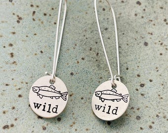 Puede incluir: Un par de pendientes de plata. Cada pendiente presenta un pequeño dije redondo con una ilustración de pez y la palabra "wild". Los pendientes tienen alambres largos y curvos.