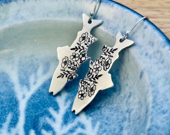 Puede incluir: Un par de pendientes plateados con forma de pez, cada uno adornado con un diseño floral negro. Los pendientes cuelgan de ganchos plateados y se exhiben en un plato con estampado azul y blanco.