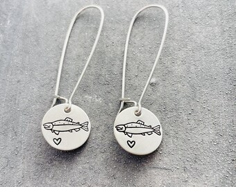 Puede incluir: Un par de pendientes de plata. Cada pendiente presenta un pequeño dije redondo con una ilustración negra de un pez y un corazón. Los pendientes tienen largos ganchos para la oreja curvados.