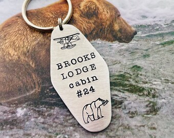 Puede incluir: Un llavero de metal plateado con forma de motel retro. La superficie está grabada con un pequeño icono de avión, el texto "BROOKS LODGE cabin #24" y un icono de oso. Una anilla metálica circular está sujeta a la parte superior del llavero.
