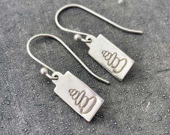 Puede incluir: Un par de pendientes de plata con un diseño rectangular. Cada etiqueta presenta un diseño grabado estilizado. Los pendientes tienen un cierre de gancho. El fondo es una superficie gris oscuro texturizada.