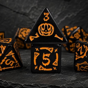 以下が含まれることがあります： ハロウィーンをテーマにしたオレンジ色の刻印が施された黒い多面体ダイスのセット。中央の4面ダイスにはジャック・オー・ランタンと数字の3が描かれ、その下の6面ダイスにはほうきのアイコンに囲まれた数字の5が刻まれています。