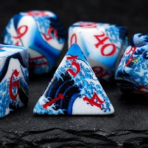 May include: A set of multi-sided polyhedral dice featuring a white base with a blue wave pattern and bold red numbers. The dice have various geometric shapes, including a four-sided triangular die in the foreground and larger rounded dice behind.