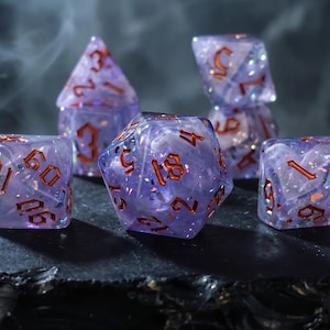 May include: Set of five purple and copper-colored polyhedral dice. The dice are translucent with glitter and copper-colored numbers. The dice are arranged on a dark surface with a blurred background.