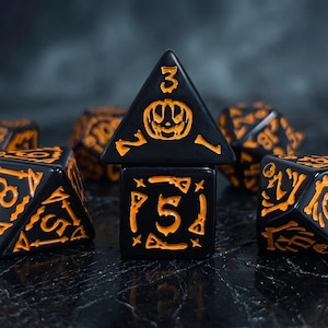 Peut inclure: Un ensemble de dés noirs et orange sur le thème d'Halloween. Les dés présentent des chiffres et des symboles orange, dont une citrouille sur un dé triangulaire. Les dés sont disposés sur une surface sombre et texturée.