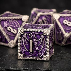 Puede incluir: Un juego de tres dados poliédricos con acabado en esmalte morado intenso y marcos de metal plateado. Cada cara muestra un motivo de dragón en relieve y números grabados, con el cubo frontal mostrando el número uno.