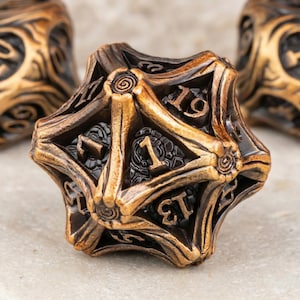 Puede incluir: Un dado de múltiples caras de color bronce con intrincadas tallas y números visibles. El dado tiene forma de estrella con una superficie detallada y texturizada. Los números son de color oscuro, contrastando con el bronce. Diseñado para juegos de mesa.
