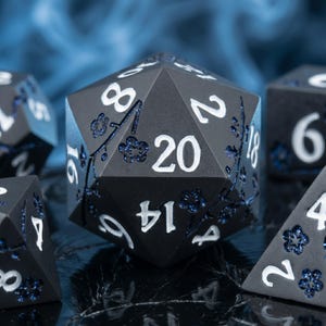 May include: A twenty-sided die featuring a matte black finish with white engraved numbers. The surface is decorated with blue glitter-filled cherry blossom branch motifs. The die displays the number 20 prominently on its front face.