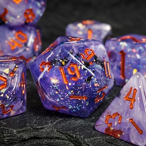 Può includere: Un set di dadi poliedrici realizzati in resina viola traslucida con nuvole bianche vorticose e scaglie di glitter iridescente incorporate. Ogni dado presenta bordi affilati e numeri incisi riempiti con un pigmento arancione metallizzato.