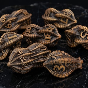 May include: A collection of bronze-colored, multi-sided dice with intricate, textured designs. Each die features gold-colored numbers. The dice are arranged on a black, marbled surface, creating a visually appealing display for tabletop gaming.
